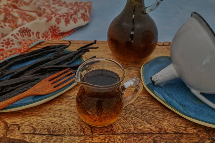 vanilla bean syrup in a small pitcher and bottle, vanilla beans and a fork on a plate, a funnel on another plate, all on a wooden board with a kitchen towel in the background