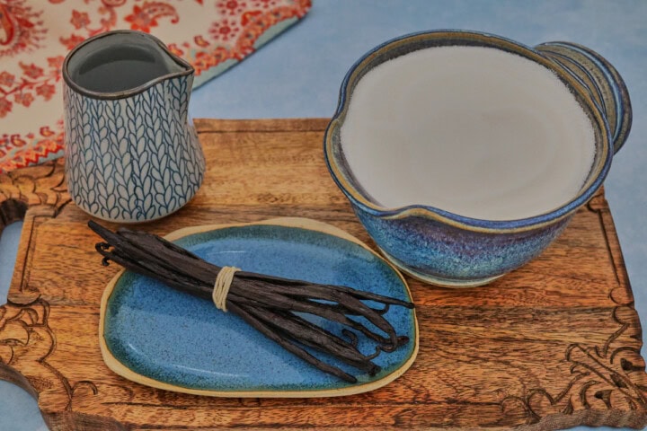 a pitcher of water, vanilla beans on a plate, and a mixing bowl of sugar on a wooden board with a kitchen towel in the background