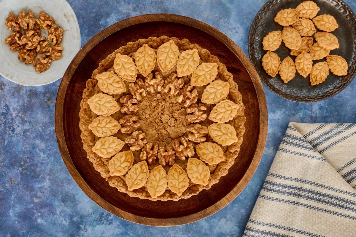a walnut tart on a cake stand with plates of walnuts and cutouts and a kitchen towel