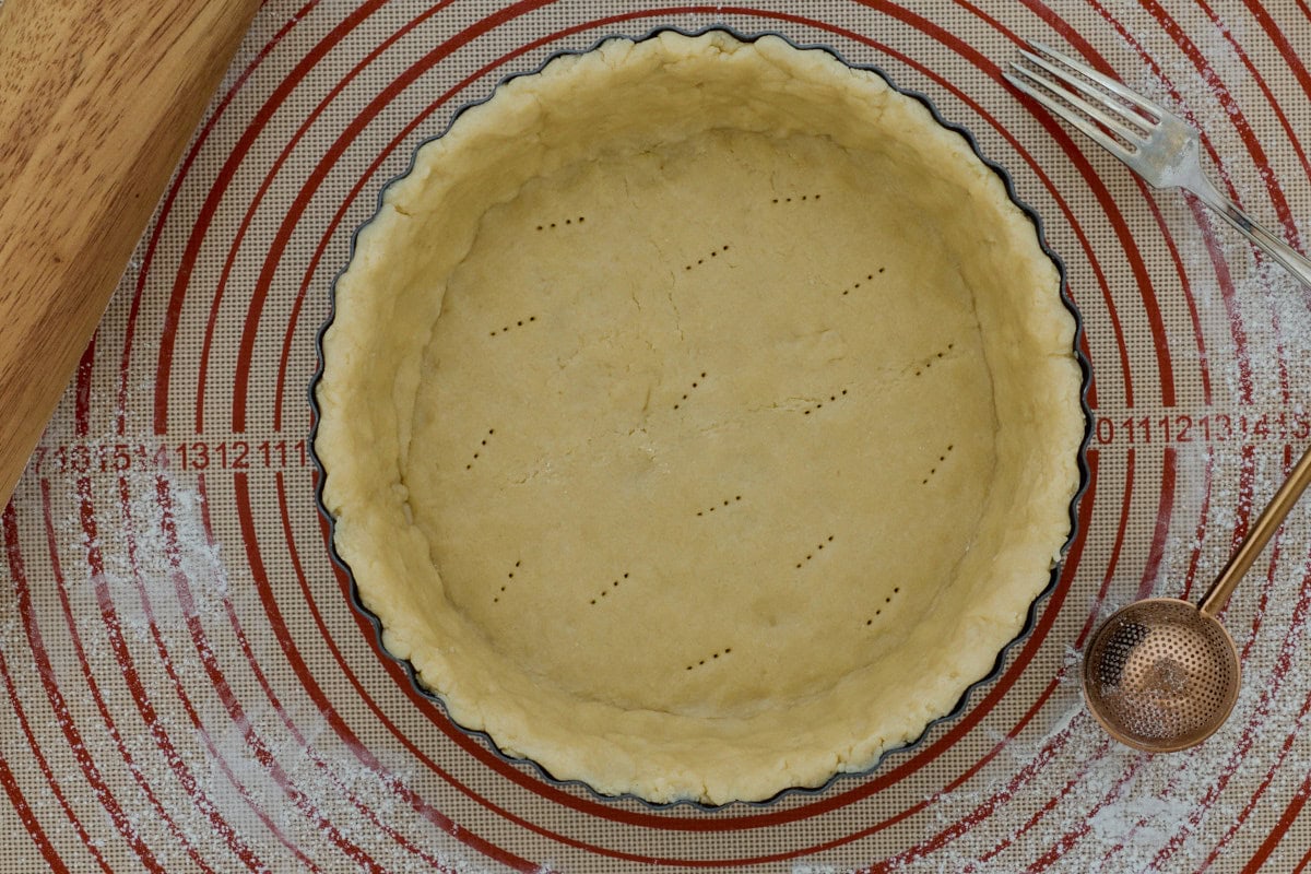docked pie crust dough in a tart pan on a silicone mat sprinkled with flour with a rolling pin, fork, and flour sprinkling wand