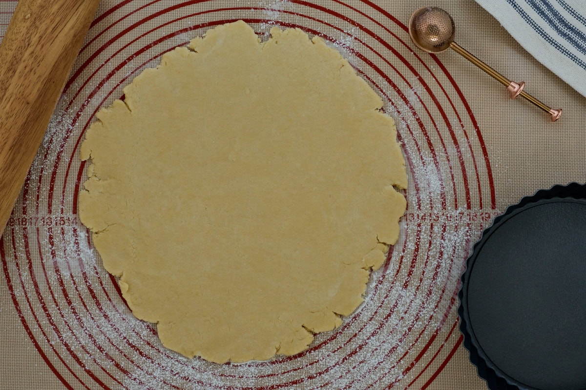 pie crust rolled out on a silicone mat with sprinkled flour, a rolling pin, flour sprinkling wand, kitchen towel, and tart pan