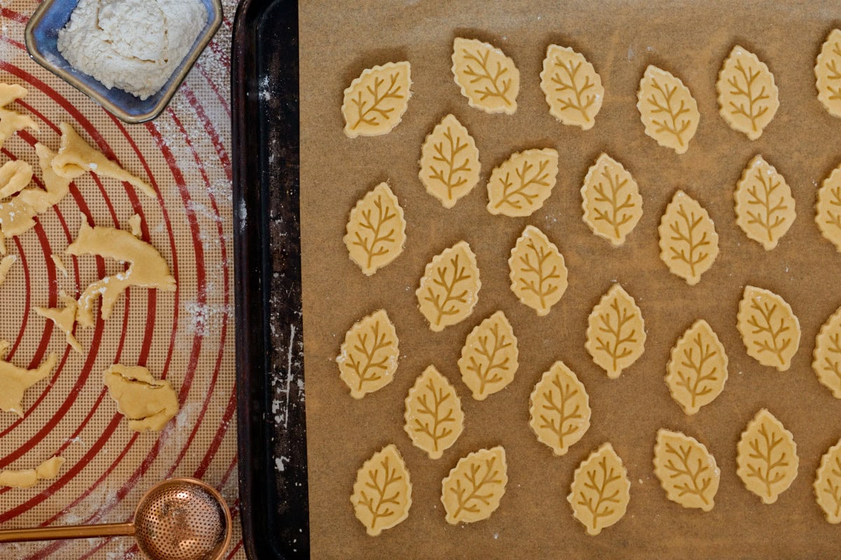 dough cutouts on parchment paper on a cookie sheet, with a silicone mat, dough scraps, flour sprinkling wand, and small bowl of flour