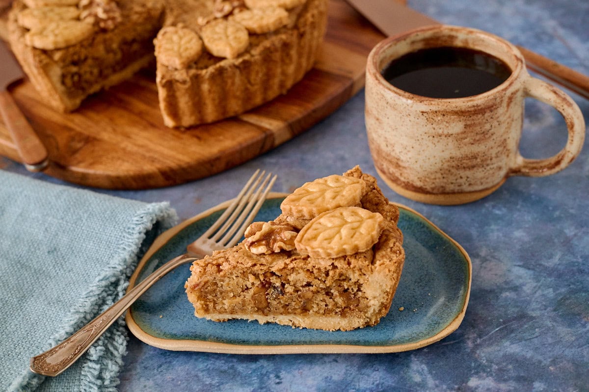 a slice of tart on a plate with a fork, napkin, cup of coffee, knife, pie server, and a walnut tart with a slice removed on a cutting board