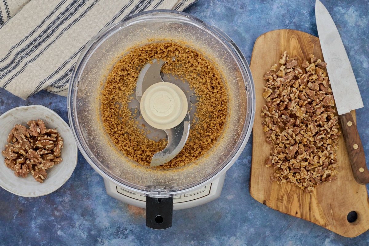 walnut halves on a bowl, walnut crumbs in a food processor, and chopped walnuts on a cutting board with a knife and a kitchen towel in the background
