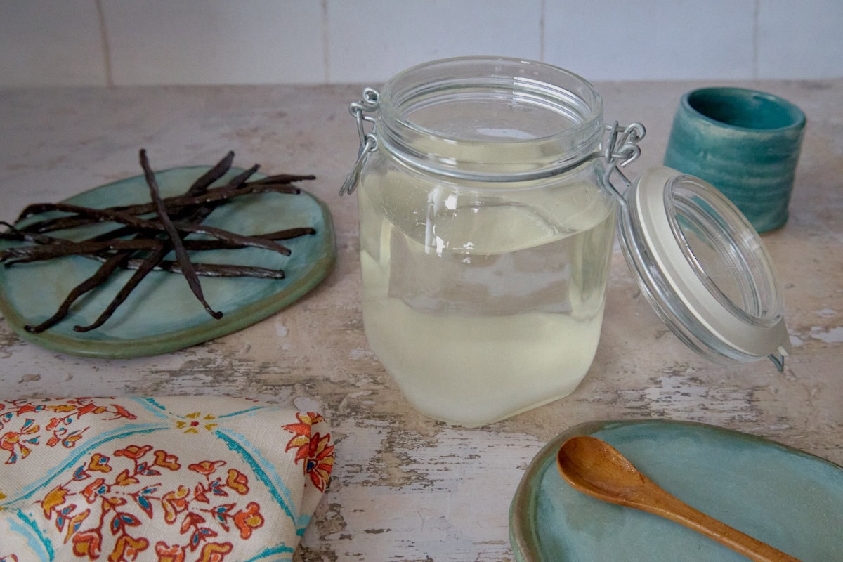 a jar with sugar and vodka, a plate of vanilla beans, a kitchen towel, a ceramic cup, and a plate with a wooden mixing spoon