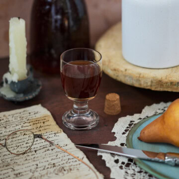 a bottle and glass of liqueur, drippy candle in a holder, glasses on handwritten documents, a cork, a tray with a vase, a doily with a plate, knife, and pear