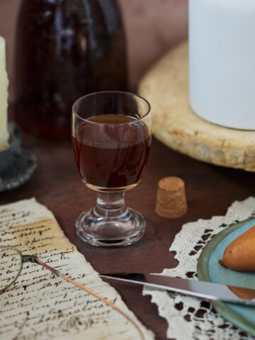 a bottle and glass of liqueur, drippy candle in a holder, glasses on handwritten documents, a cork, a tray with a vase, a doily with a plate, knife, and pear