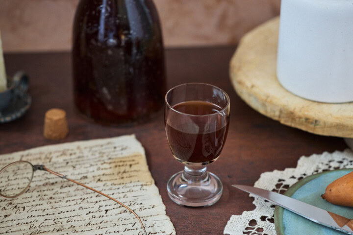 a glass and bottle of liqueur, a candle in a holder, glasses on handwritten documents, a cork, a vase on a tray, and a doily, plate, knife, and pear
