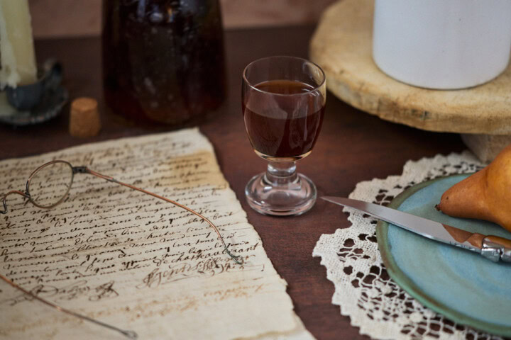 a glass and bottle of liqueur, candle in a holder, cork, glasses on handwritten documents, a tray with a vase, a lace doily with a plate, knife, and pear