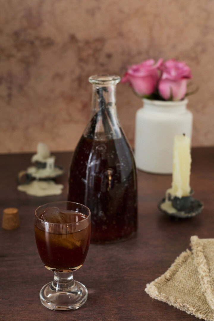 a bottle and glass of liqueur, drippy candles in candle holders, a cork, vase of roses, and a napkin