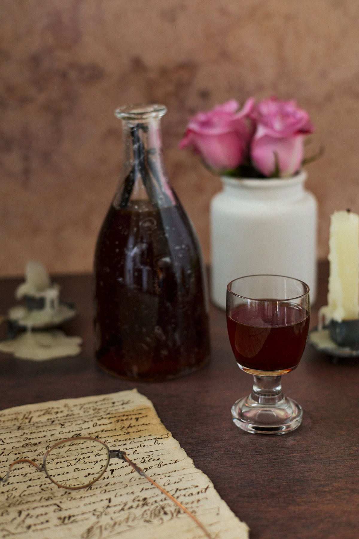a bottle and glass of liqueur, drippy candles in candle holders, a handwritten document with glasses, and a vase of roses