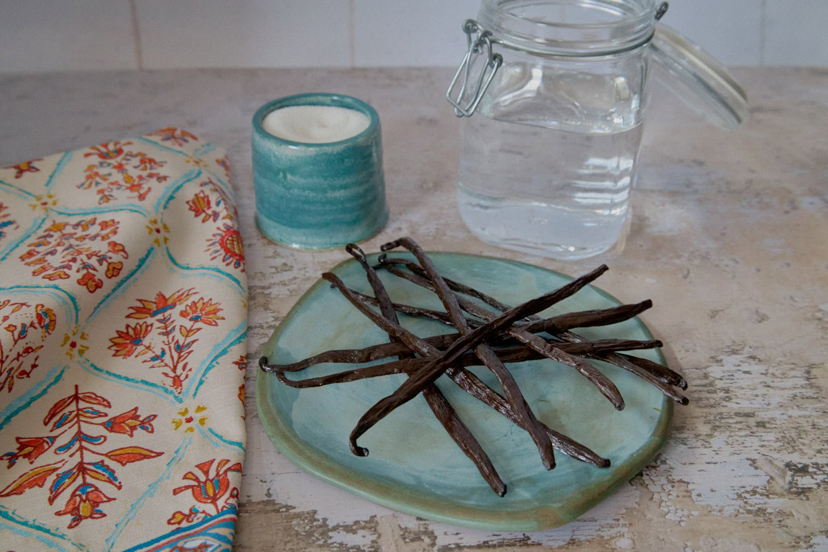 a plate of vanilla beans, kitchen towel, cup of sugar, and jar of vodka