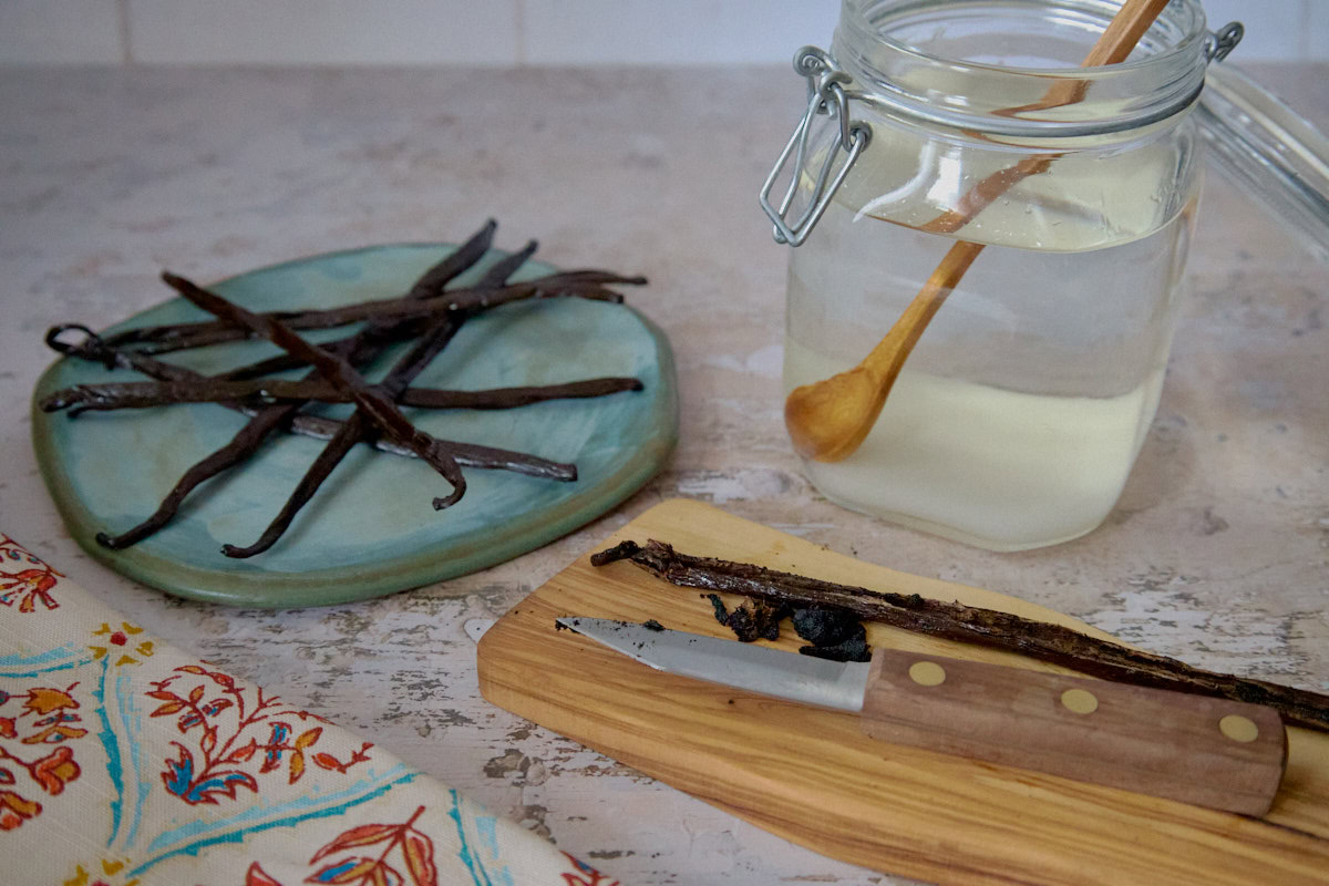 a plate of vanilla beans, jar of sugar and vodka with a mixing spoon, cutting board with knife, vanilla bean, and vanilla seeds, and a kitchen towel