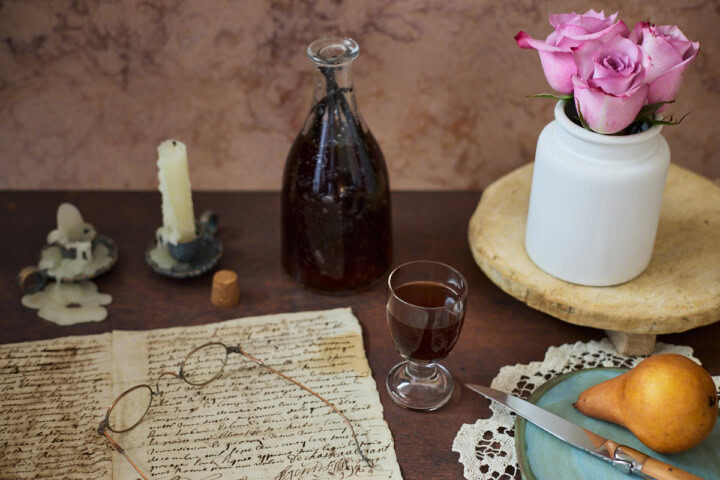 a glass of liqueur, handwritten documents, glasses, a candle in a holder, a bottle of liqueur and a cork, a tray with a vase of roses, a lace doily, and a plate with a knife and pear