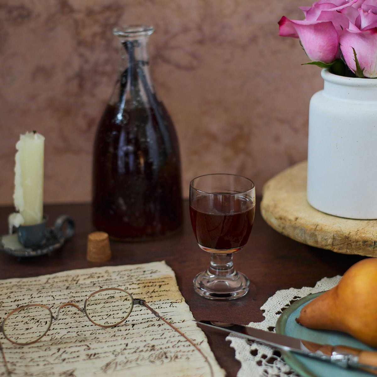 a glass of liqueur, handwritten documents, glasses, a candle in a holder, a bottle of liqueur and a cork, a tray with a vase of roses, a lace doily, and a plate with a knife and pear
