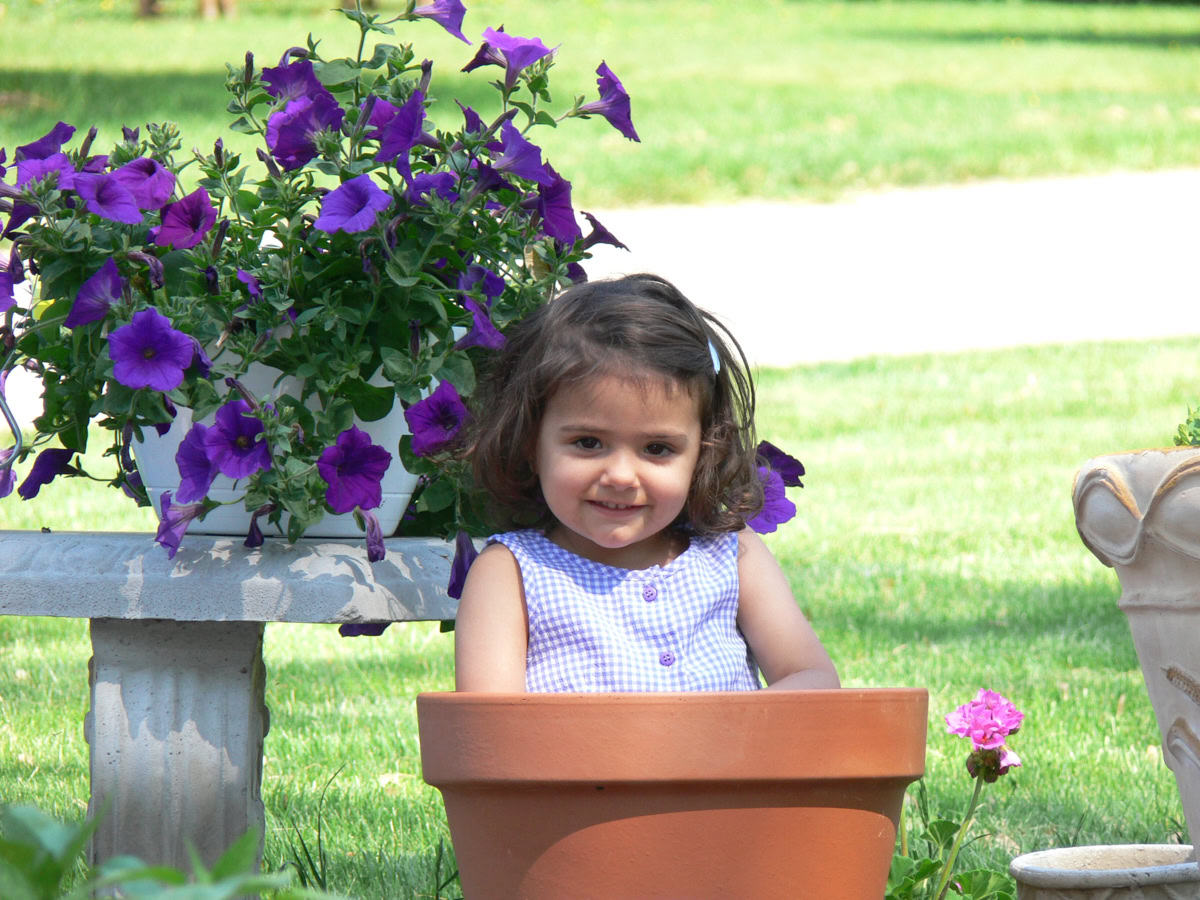 a young girl sitting in a large terra cotta pot in a garden next to a bench with potted flowers on it