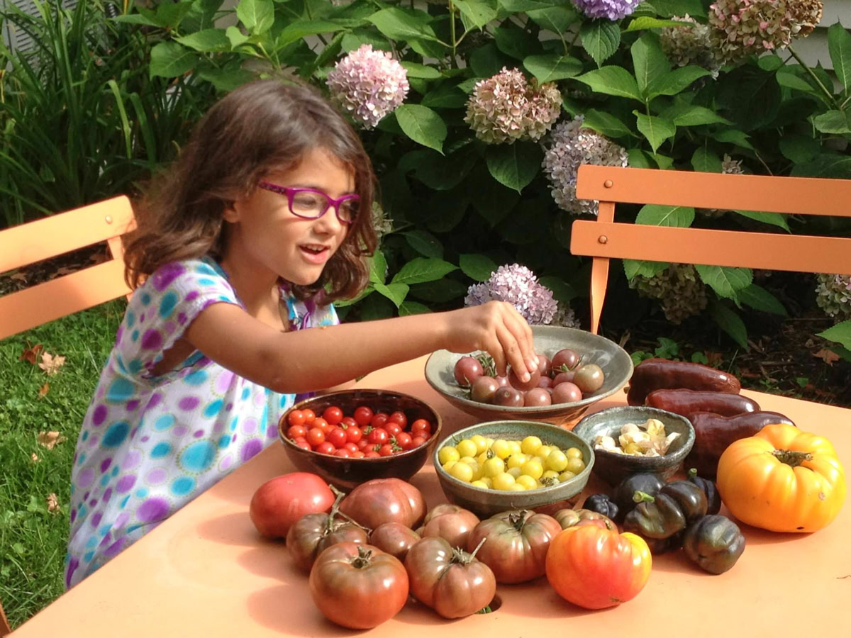a young girl tasting freshly-picked tomatoes and peppers in bowls on an outdoor table