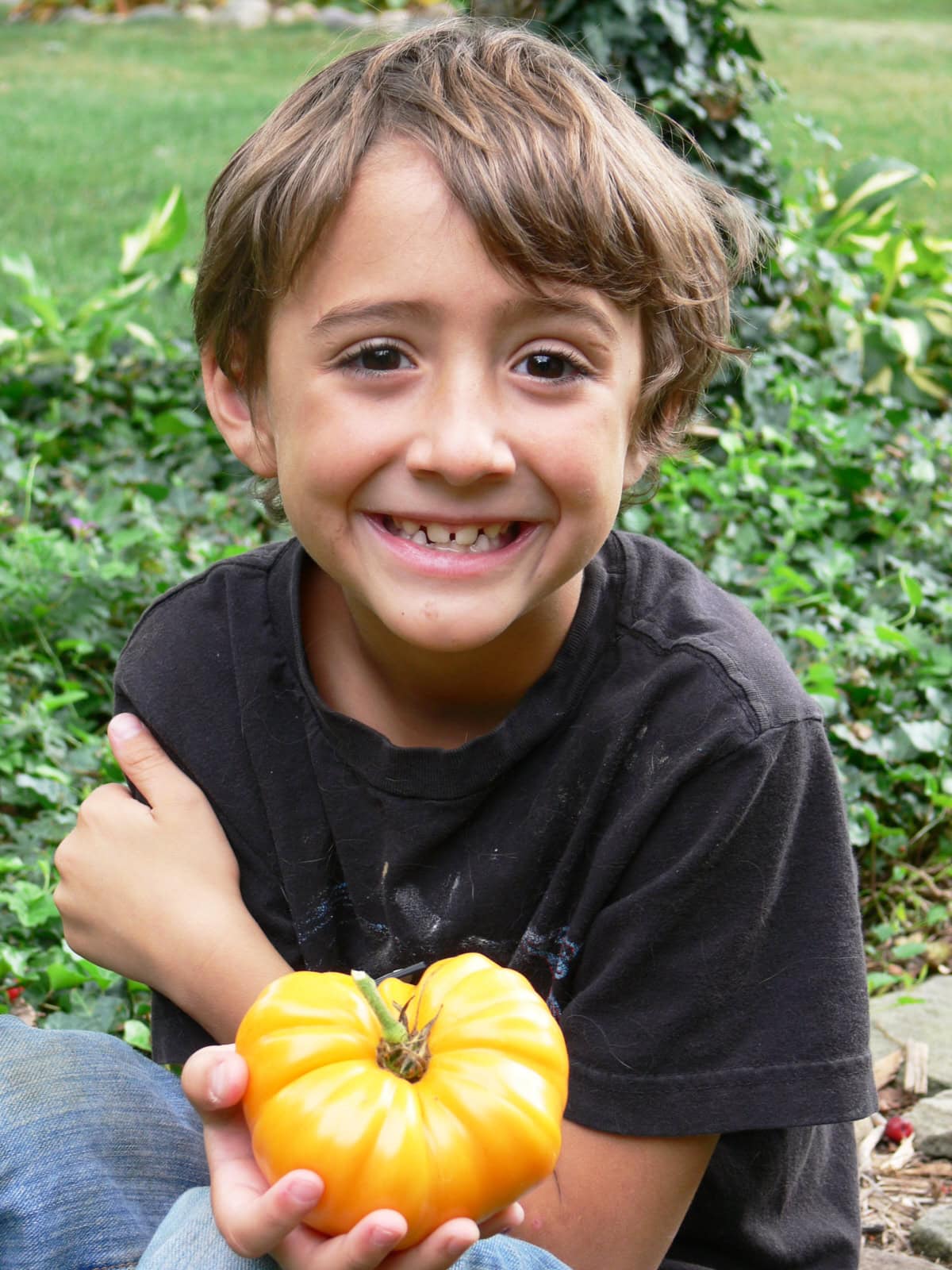 a boy sitting on the ground, holding a yellow tomato, and smiling