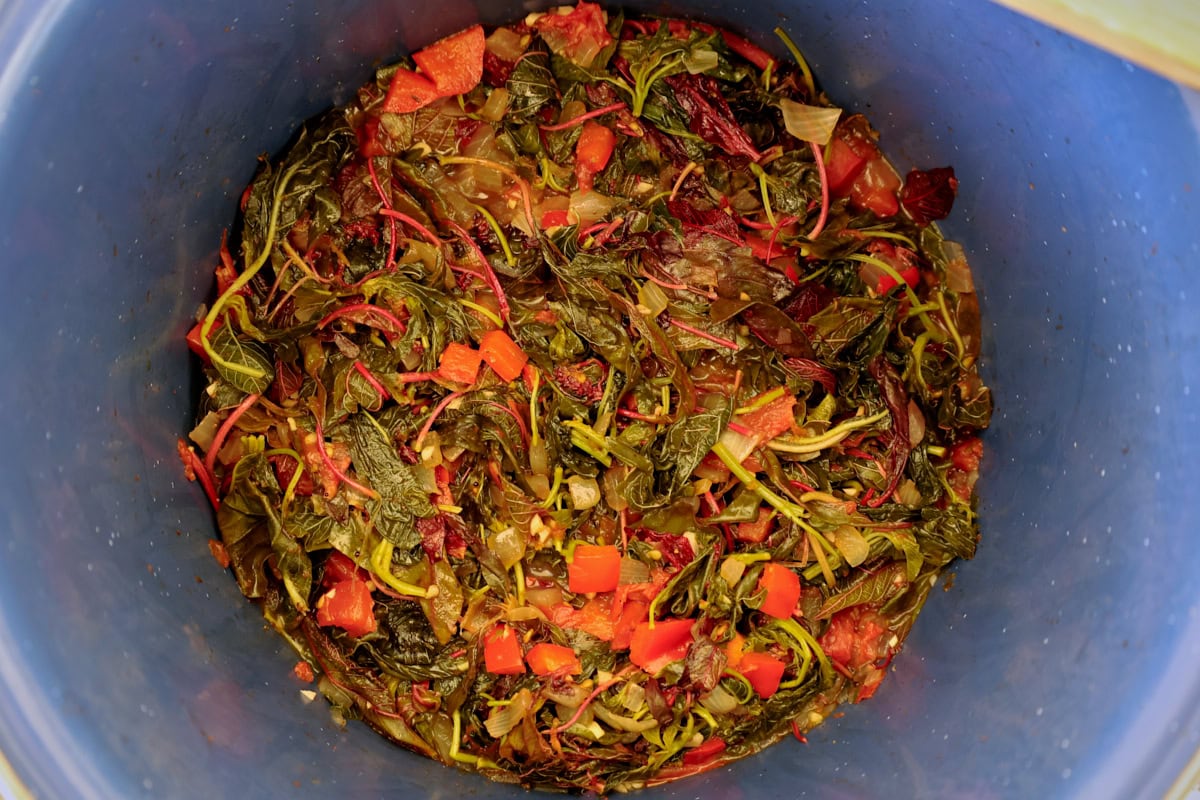 cooked amaranth greens with onions and tomatoes in a blue pot, with a wooden mixing spoon