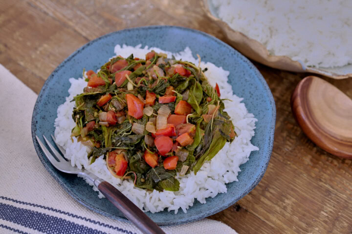 cooked amaranth greens with onions and tomatoes over rice in a bowl with a fork, with a kitchen towel, wooden spoon, and bowl of rice