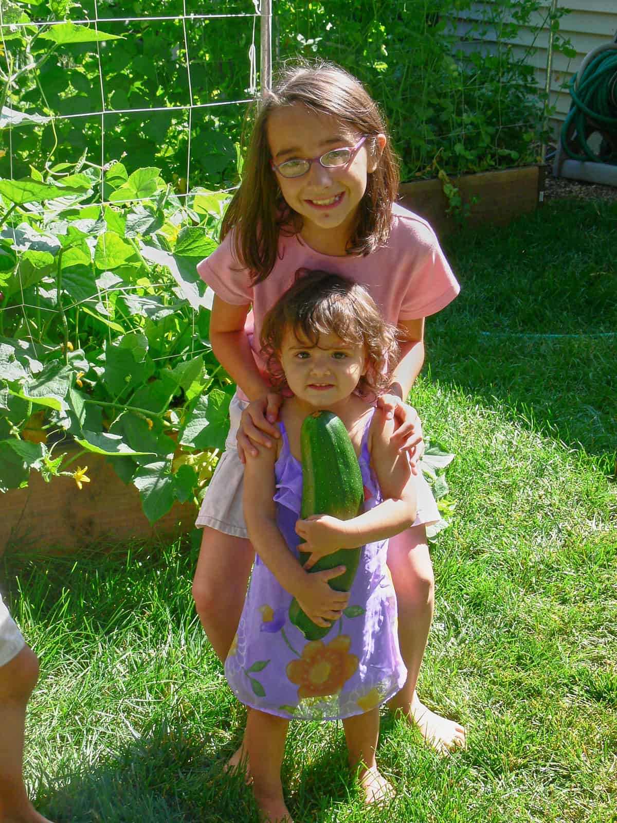 two girls standing in front of raised bed gardens, one in front of the other; the youngest in front holds a large zucchini