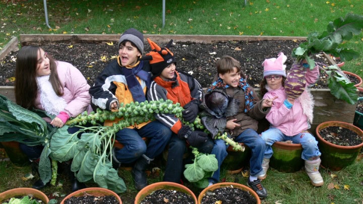 5 kids sitting in planters next to a raised garden bed, holding freshly-harvested fall crops