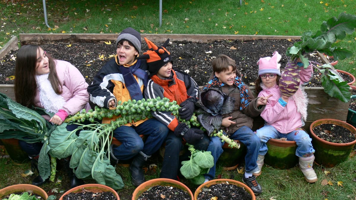 5 kids sitting in planters next to a raised garden bed, holding freshly-harvested fall crops