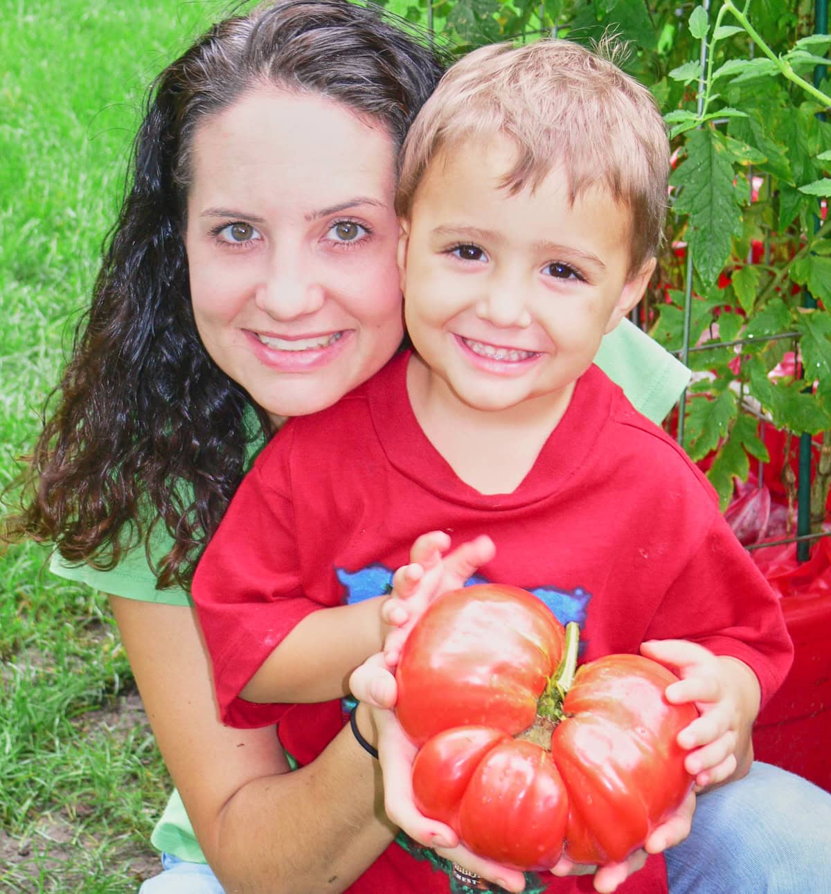 a woman with a young boy standing in front of her, both holding a large red tomato and smiling
