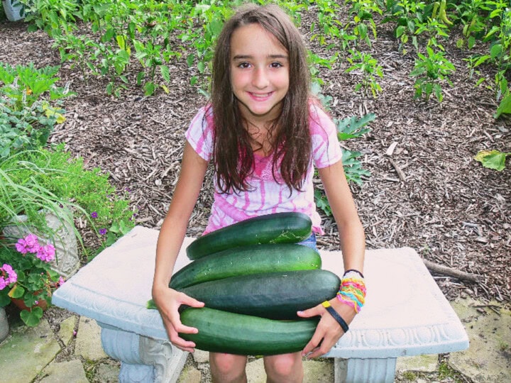 a girl on a bench in front of a garden holding some zucchini