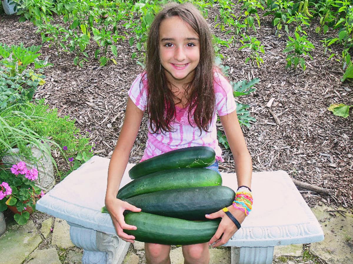 a girl on a bench in front of a garden holding some zucchini