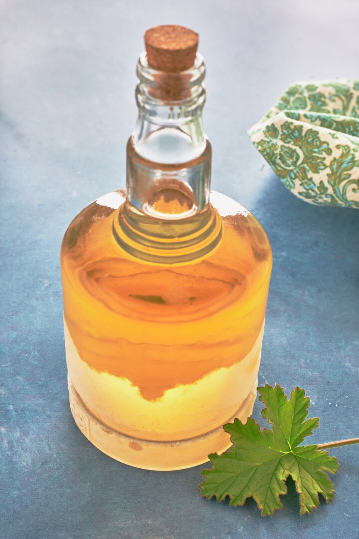attar of roses scented geranium liqueur in a bottle with a cork with a fresh leaf in front and a napkin