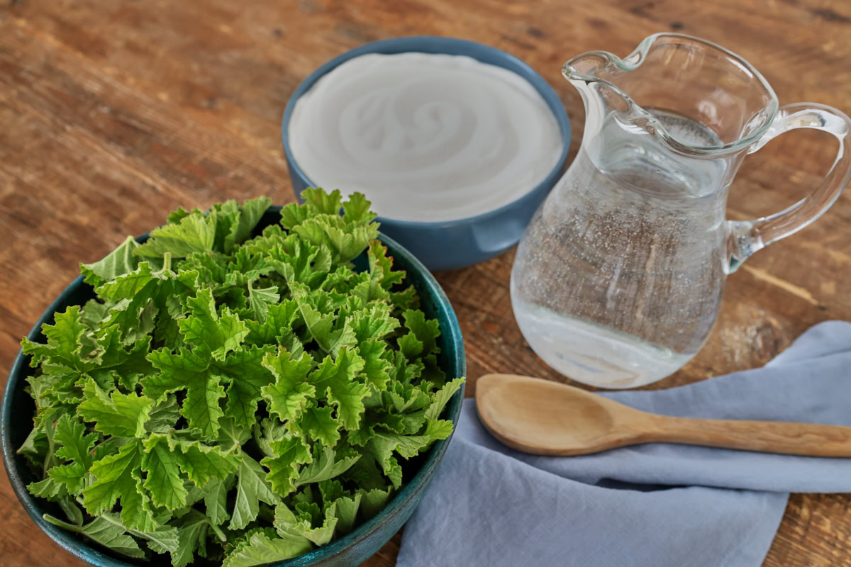 scented geranium leaves in a bowl, sugar in a bowl, a napkin, wooden spoon, and a pitcher of clear liquid