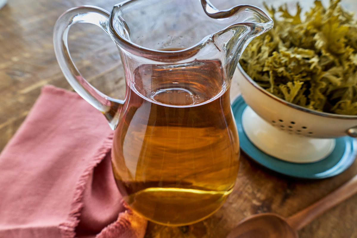 strained leaves in a colander over a plate, a napkin, wooden spoon, and amber liquid in a pitcher
