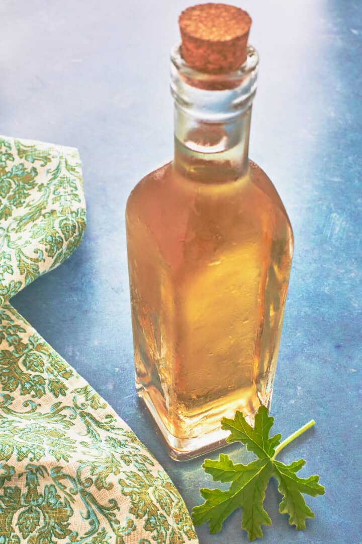 true rose scented geranium liqueur in a bottle with a cork with a fresh leaf in front and a napkin