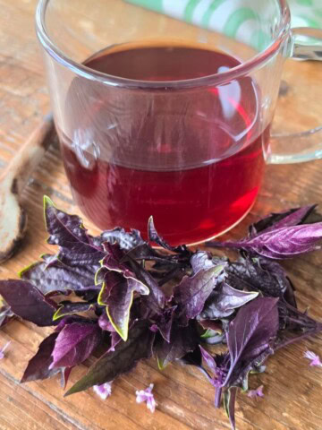 a cup of basil tea on a wooden table with fresh basil leaves and flowers in front, a spoon alongside, and a kitchen towel in the back