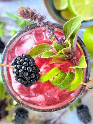 an overhead view of a blackberry basil lime rickey cocktail or mocktail, with garnishes in the drink and in the background on the table